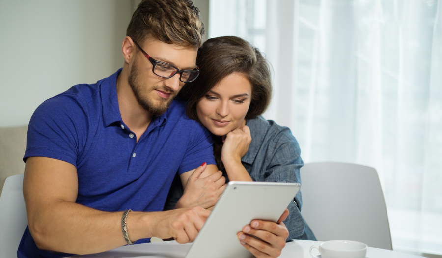 A man wearing glasses sits next to a woman on a couch and they look at a tablet